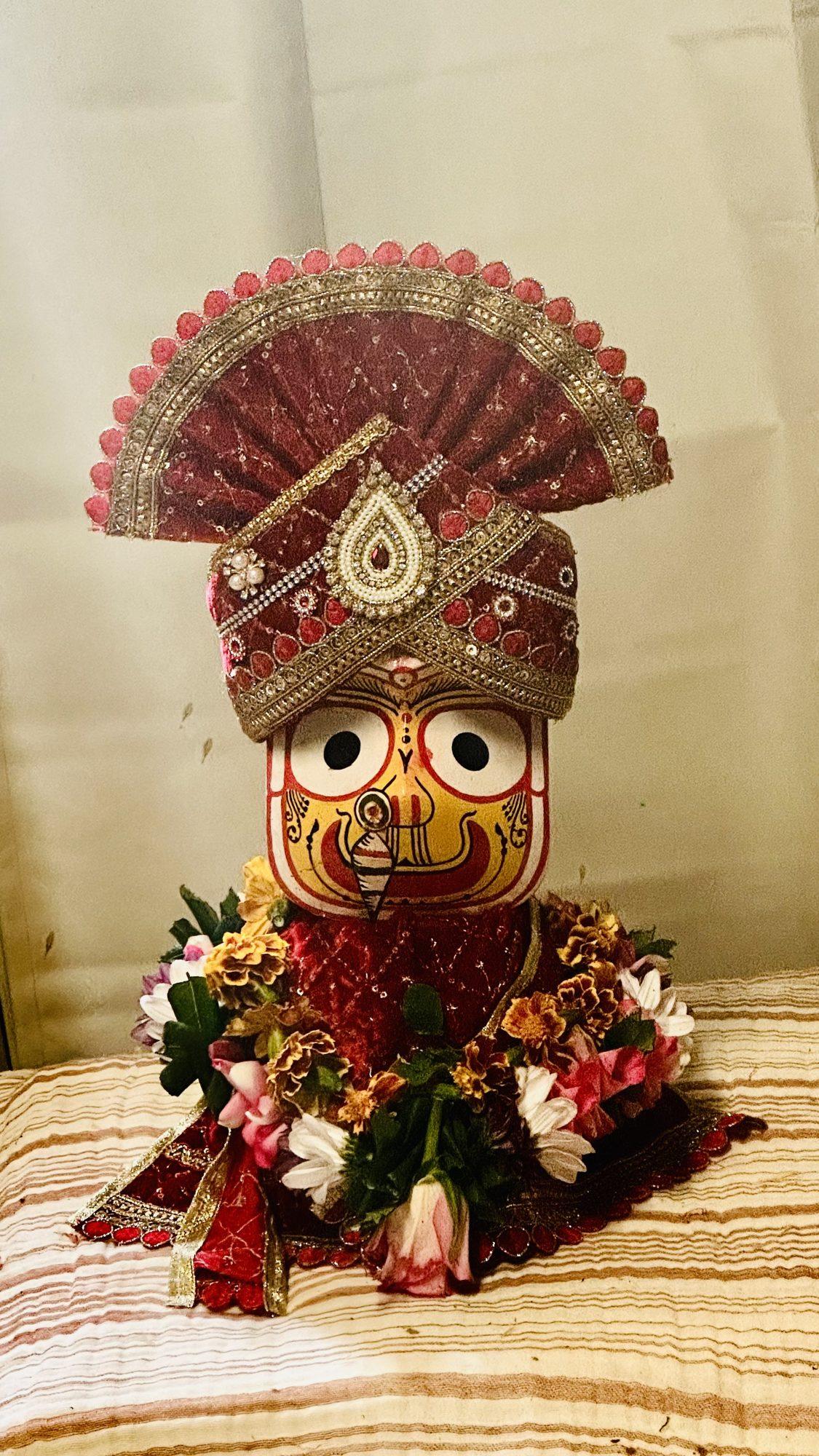 A Jagannath idol decorated with a red and gold headdress and a garland of fresh flowers