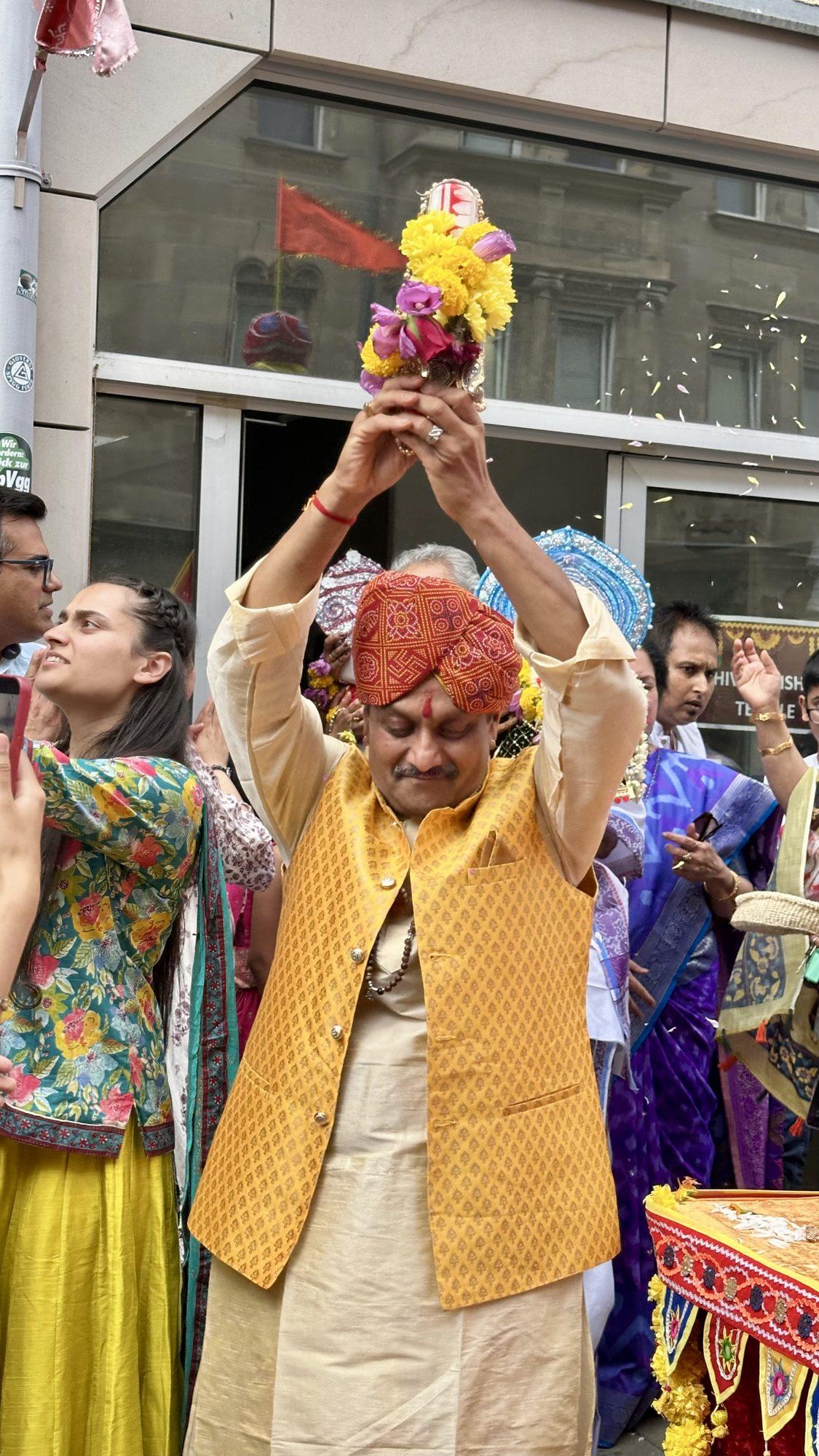 A man in a yellow waistcoat holds flowers high during the Rath Yatra procession as flower petals fall around him