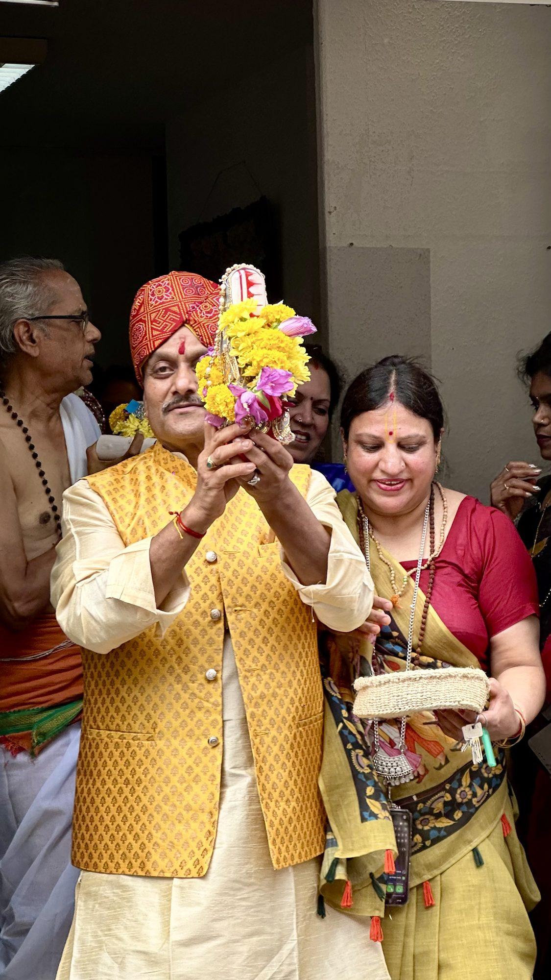 A man in a yellow waistcoat and orange turban raising a garland of marigolds while devotees look on